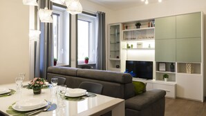 Un'elegante zona pranzo con un tavolo bianco apparecchiato per due, un divano grigio e una libreria a parete con oggetti decorativi.