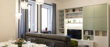 Un comedor moderno con una mesa blanca dispuesta para dos personas, un sofá gris y una estantería empotrada con adornos.