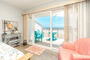 A room with a bed, a desk, a chair, a small table, and a view of the beach through a sliding glass door.