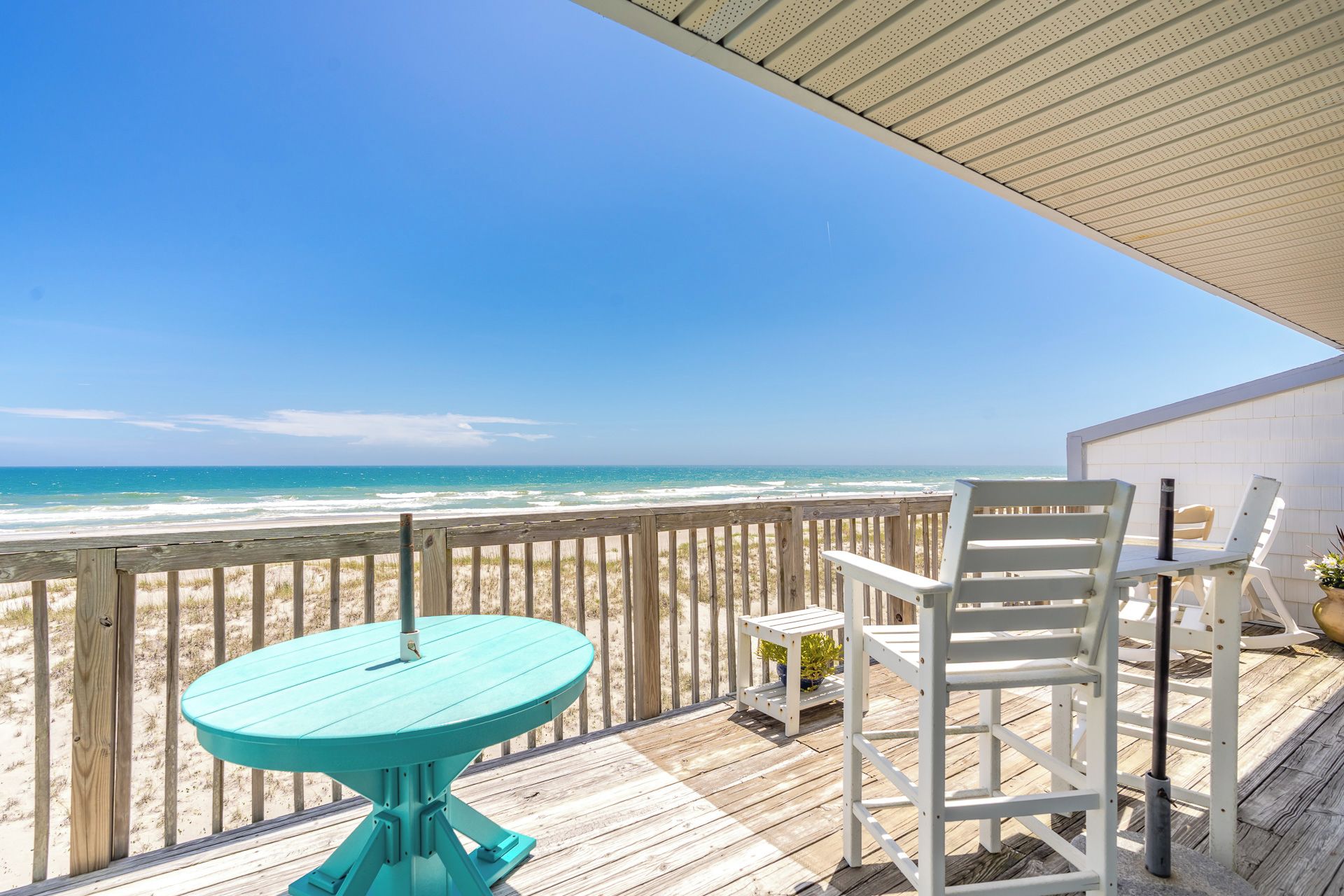 Une terrasse en bois avec une table turquoise et des chaises blanches, donnant sur une plage.