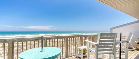 Une terrasse en bois avec une table turquoise et des chaises blanches, donnant sur une plage.