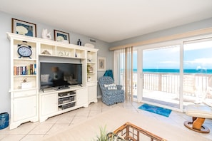 A living room with a TV, shelves with books, a chair, and a balcony with a view of the ocean.