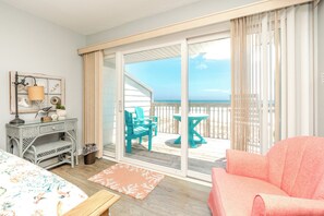 A room with a bed, a desk, a chair, a table, and a view of the beach through a sliding door. - Ocean Potion - Oceanfront Queen's Grant Condo with Amazing Views! (Topsail Beach)