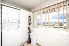 Ett tvättrum med en tvättmaskin och en torktumlare som man öppnar framifrån, en sopkorg och ett fönster med persienner.