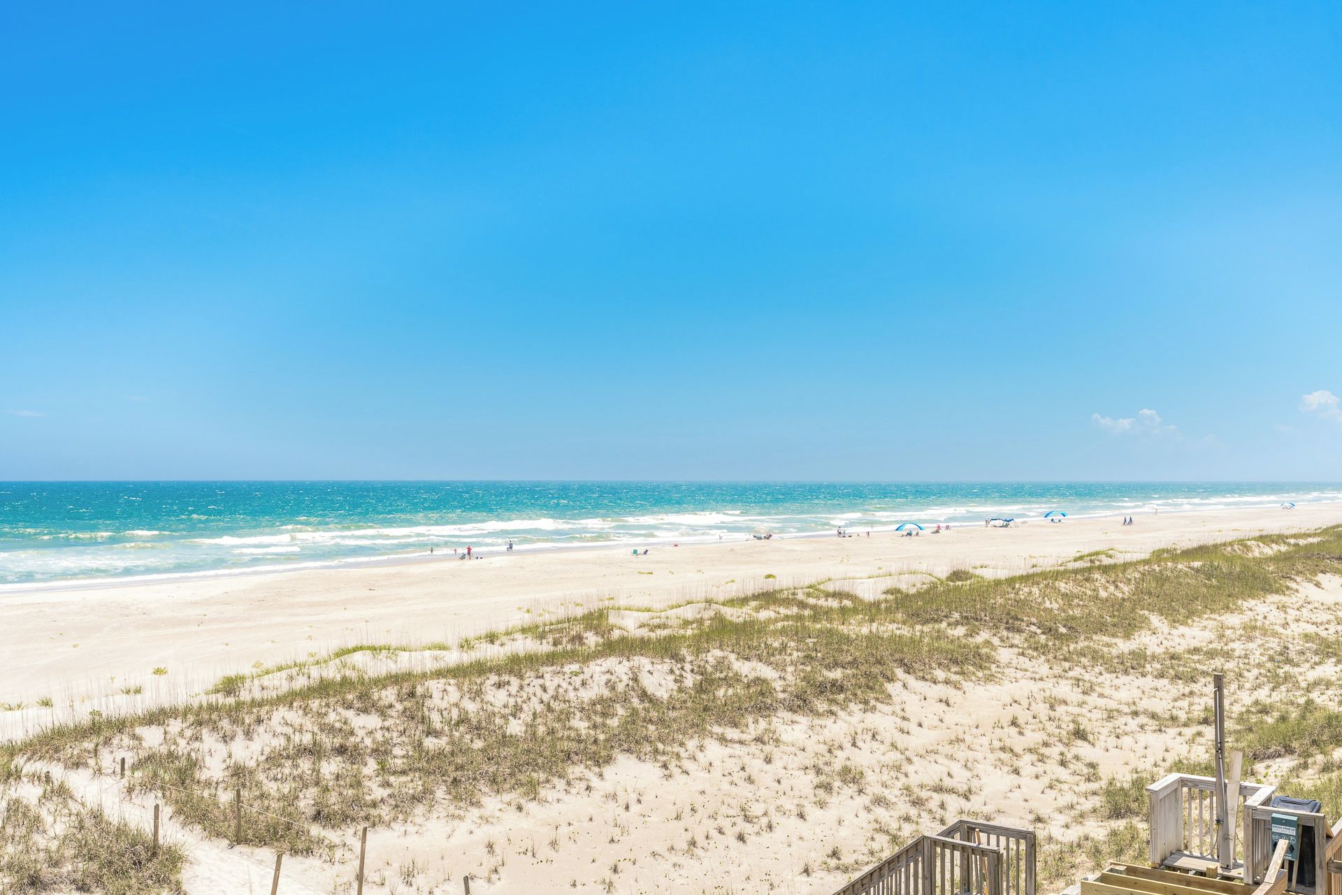 Une plage avec une promenade en bois qui mène à l’eau, des dunes de sable et un ciel bleu clair.