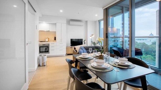 A modern apartment with a dining area, kitchen, and large windows offering a city view. - City Living At Best Location In Melbourne (Melbourne)