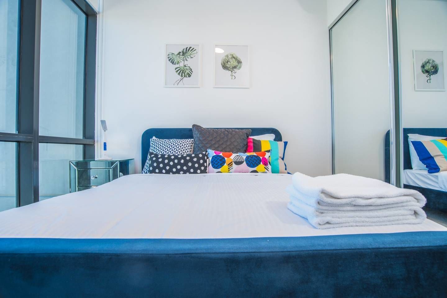 A modern hotel room with a large bed, colorful pillows, a glass side table, and two wall art pieces.