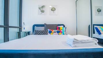 A modern hotel room with a large bed, colorful pillows, a glass side table, and two wall art pieces.
