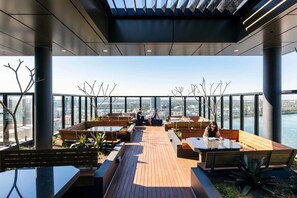 Een terras op het dak met houten zitplaatsen, een glazen balustrade en uitzicht op de skyline.