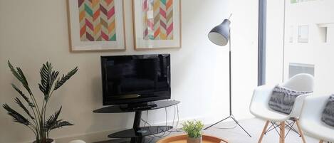 A modern living room with a flat-screen TV, two framed artworks, a potted plant, a round wooden coffee table, a floor lamp, and a white armchair.