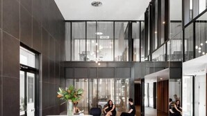 A modern lobby with a high ceiling, dark walls, and large windows. There are seating areas with people and a plant. - Luxury 1 Bedroom 1 Study With 1 Parking (Wentworth Point)