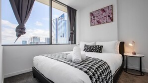 A hotel room with a large bed, a view of the cityscape, a painting on the wall, and a small table with a lamp. - New Renovated & Cozy Apt Closes To Southern Cross (Melbourne)