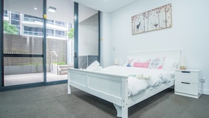 Une chambre moderne avec un lit blanc, une table de chevet et une grande fenêtre donnant sur un bâtiment et sur la verdure environnante.
