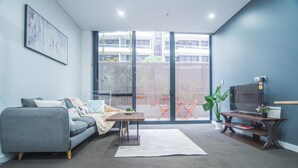 Un salon moderne avec un canapé gris, une table basse, un téléviseur posé sur un meuble en bois et une grande porte-fenêtre coulissante donnant sur une terrasse extérieure.