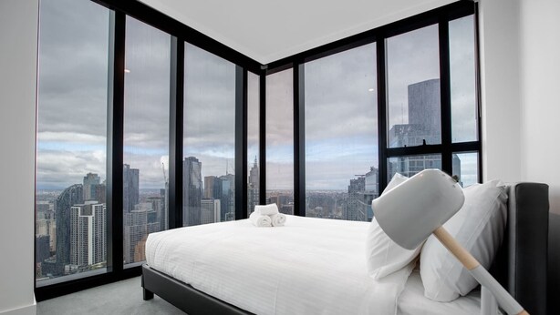 Une chambre d’hôtel moderne dotée d’un grand lit, d’une table de chevet et offrant une vue sur le paysage urbain grâce à de grandes fenêtres du sol au plafond.