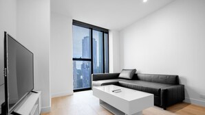 Un salon moderne doté d’un grand téléviseur à écran plat, d’un canapé gris, d’une table basse blanche et offrant une vue sur les immeubles de la ville par le biais d’une grande fenêtre.