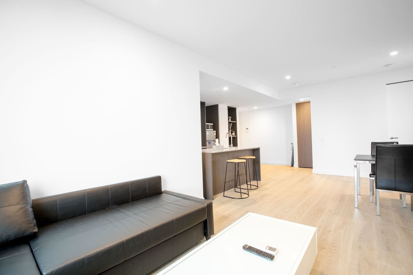 Een moderne woonkamer met een donker leren bank, een witte salontafel en een keuken met een bar en barkrukken.