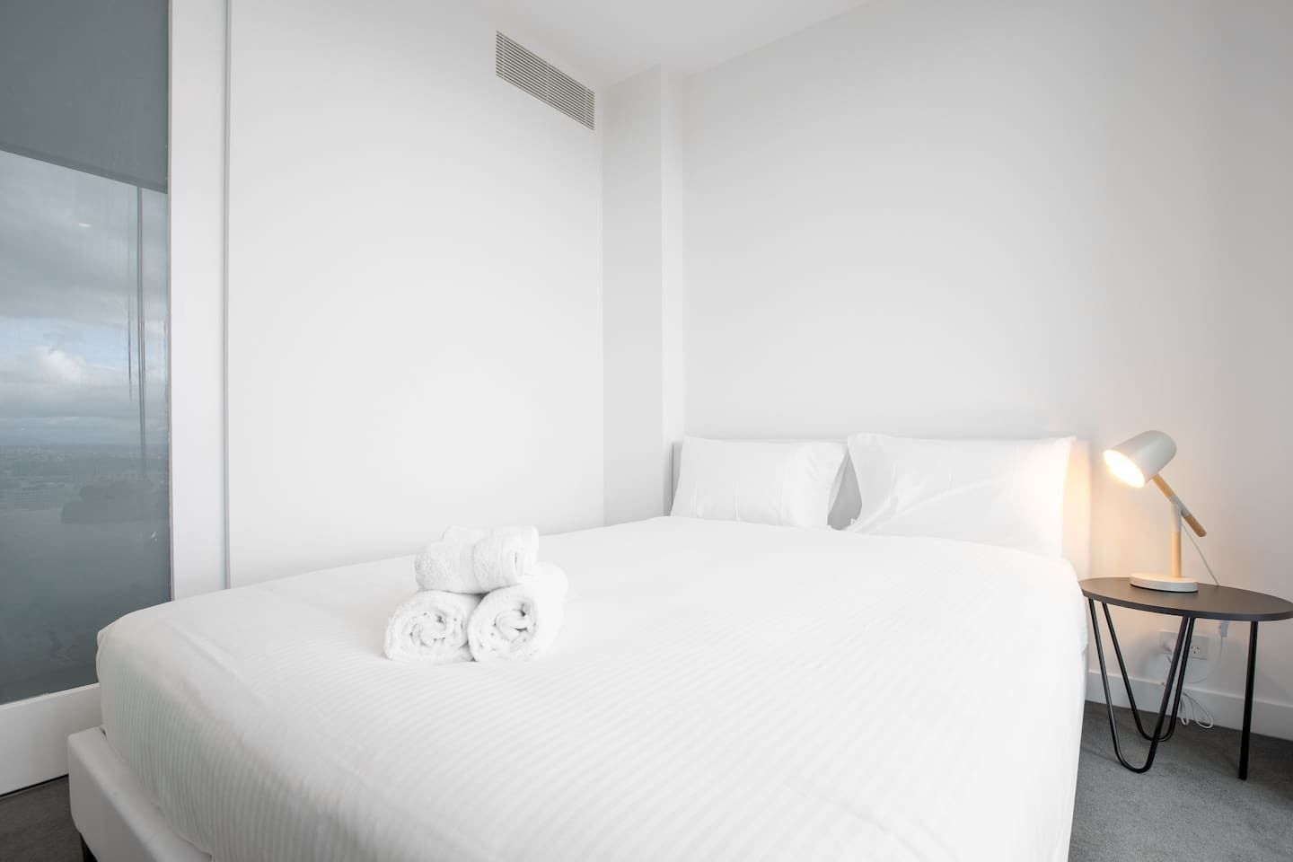 A modern hotel room with a large bed, white linens, a bedside lamp, and a view through a large window.