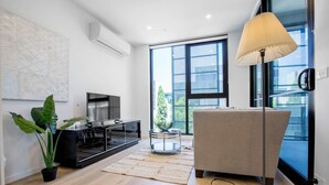 A modern living room with a flat-screen TV, a beige sofa, a floor lamp, and a large window offering a view of buildings and trees. - Stunning Bright Apartment At Hawthron/Glenferrie Station (Hawthorn)