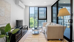 A modern living room with a flat-screen TV, a beige sofa, a coffee table, and large windows offering a view of the city. - Stunning Bright Apartment At Hawthron/Glenferrie Station (Hawthorn)