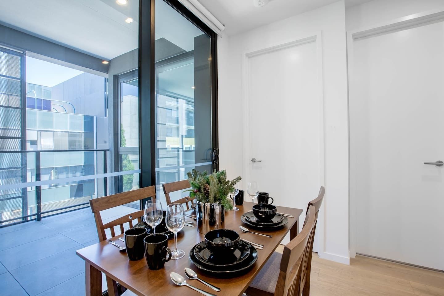 A dining area with a table set for four, including black plates, bowls, and cutlery. There is a vase with greenery on the table.