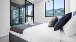 A modern bedroom with a large bed, a window with a view of buildings, and a glass door leading to an outdoor area. - Stunning Bright Apartment At Hawthron/Glenferrie Station (Hawthorn)