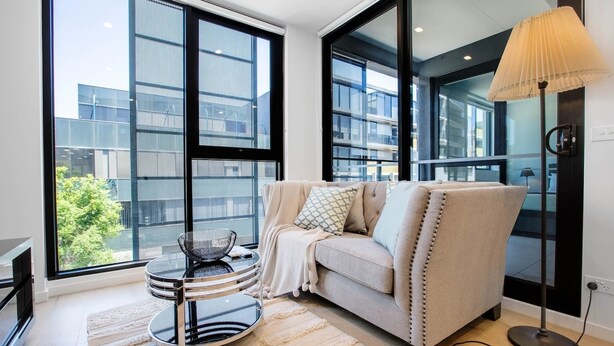 A modern living room with a sofa, a round table, and a floor lamp. Large windows offer views of buildings and greenery. - Stunning Bright Apartment At Hawthron/Glenferrie Station (Hawthorn)