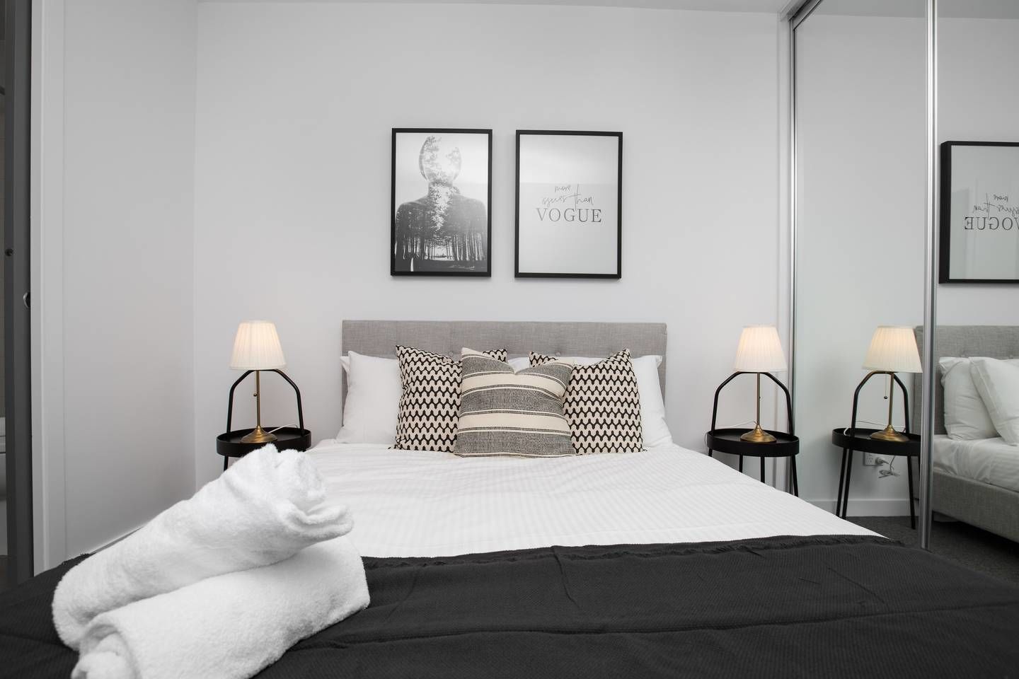 Habitació d'hotel moderna amb un llit gran, dues taules de nit amb làmpades, un capçal de llit gris i dos quadres al mur.