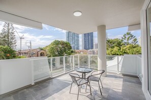 Ein Balkon mit Glasgeländer, einem Tisch und zwei Stühlen sowie Blick auf hohe Gebäude und Bäume.