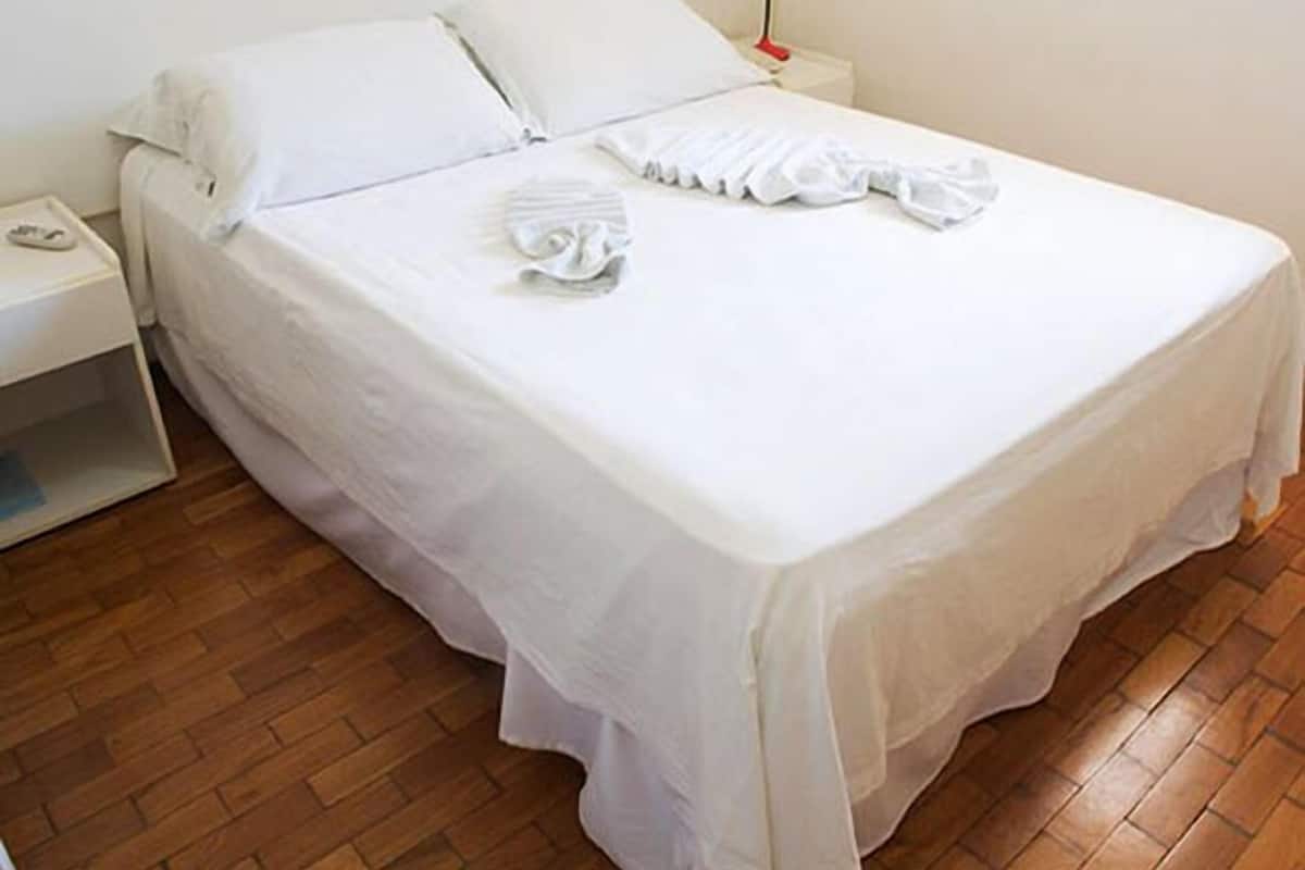 Uma cama bem arrumada com lençóis brancos, uma mesa de cabeceira com abajur e piso de madeira.