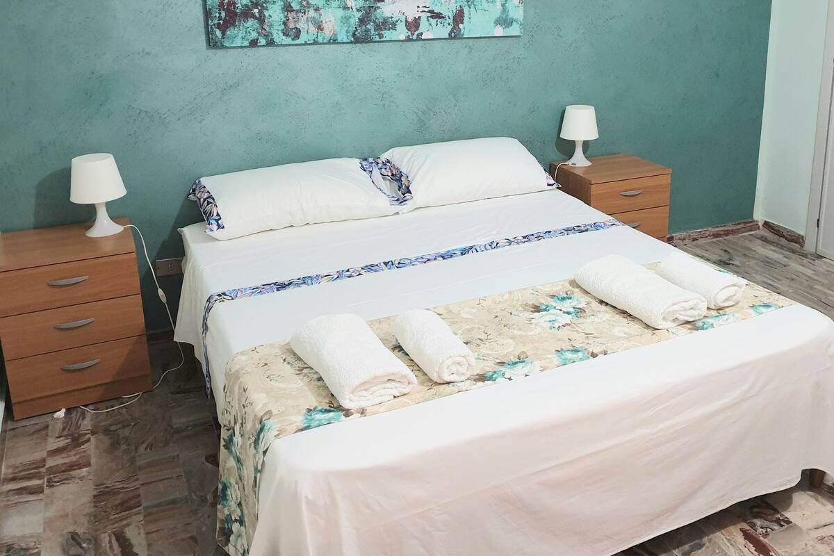 Cama bem arrumada com lençóis brancos e colcha florida, duas luminárias de cabeceira e mesa de cabeceira de madeira com gavetas.