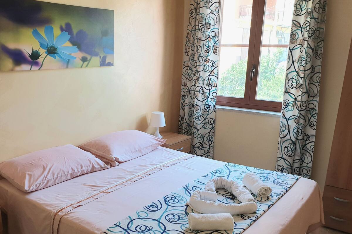 Cama bem arrumada com roupa de cama rosa, abajur e quadro floral na parede.