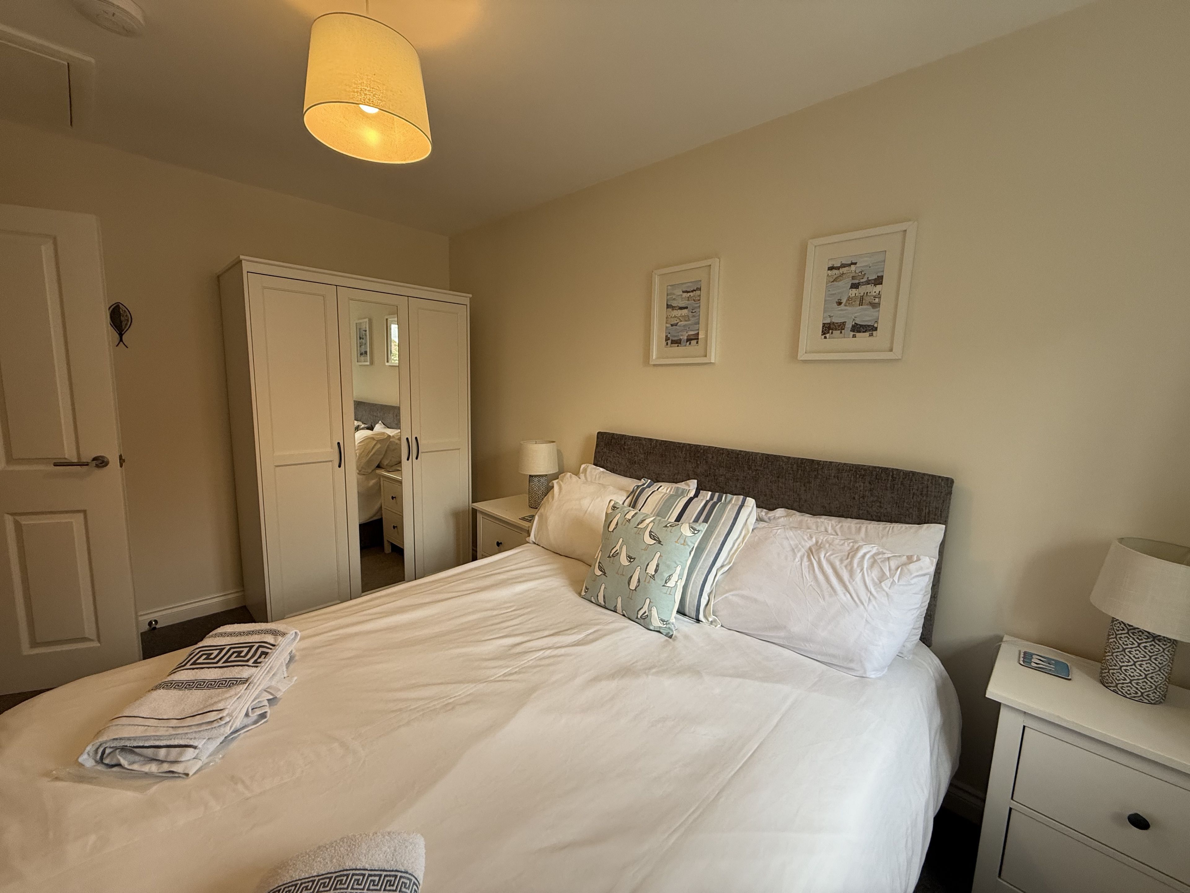 A neatly made bed with pillows and a blanket, a bedside table with a lamp, a wardrobe, and two framed pictures on the wall.