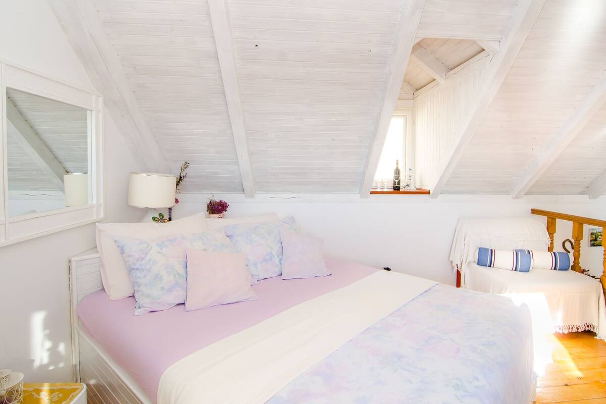 A bedroom with a sloped ceiling, a bed with pastel bedding, a bedside table with a lamp, and a window with curtains.