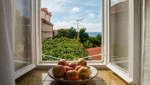 Un saladier rempli de fruits est posé sur un rebord de fenêtre, d’où l’on aperçoit un bâtiment et de la verdure à l’extérieur.