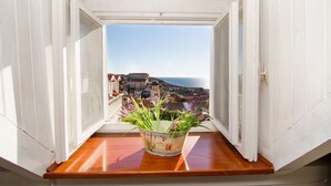 Une fenêtre donnant sur une ville côtière et un balcon agrémenté d’un pot de fleurs.