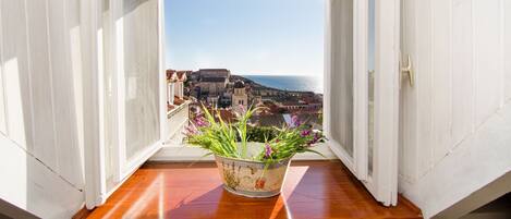 A window with a view of a coastal town and a balcony with a potted plant.