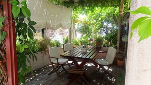 Une terrasse extérieure avec une table en bois et quatre chaises, entourée d’une végétation luxuriante et d’un auvent blanc.