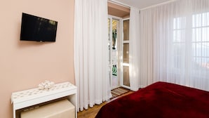 A hotel room with a TV mounted on the wall, a desk with a floral-patterned tablecloth, a red bedspread, and a sliding glass door leading to a balcony. - Rooms Lydia - Adults Only (Dubrovnik)