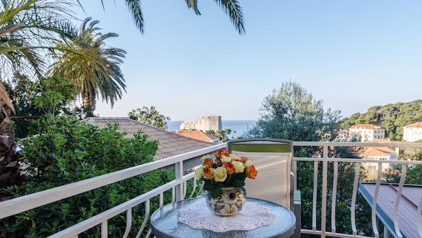 A balcony with a table holding a vase of flowers, surrounded by greenery and overlooking a coastal town with buildings and hills. - Rooms Lydia - Adults Only (Dubrovnik)