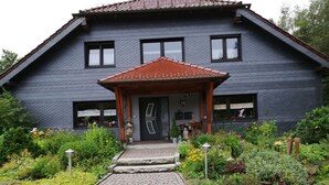 Een tweelaags huis met een donkere buitenkant en een dak van rode tegels. Er is een veranda met een klein dak en treden die naar de deur leiden.