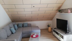 Een moderne woonkamer met een grijze hoekbank, een flatscreen televisie en een houten plafond.