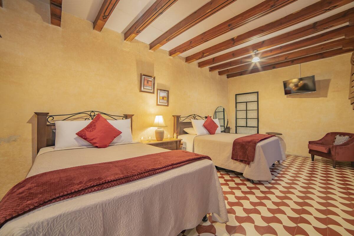 Quarto de hotel com duas camas, vigas de madeira no teto, piso estampado e televisão na parede.