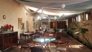 Un salon chaleureux avec une table ronde en verre, des fauteuils en osier et un canapé. Les murs sont ornés d’objets décoratifs et une grande plante se trouve près de la fenêtre.