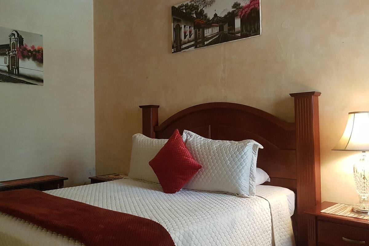 Cama bem arrumada com travesseiro vermelho, cabeceira de madeira e mesa de cabeceira com abajur.