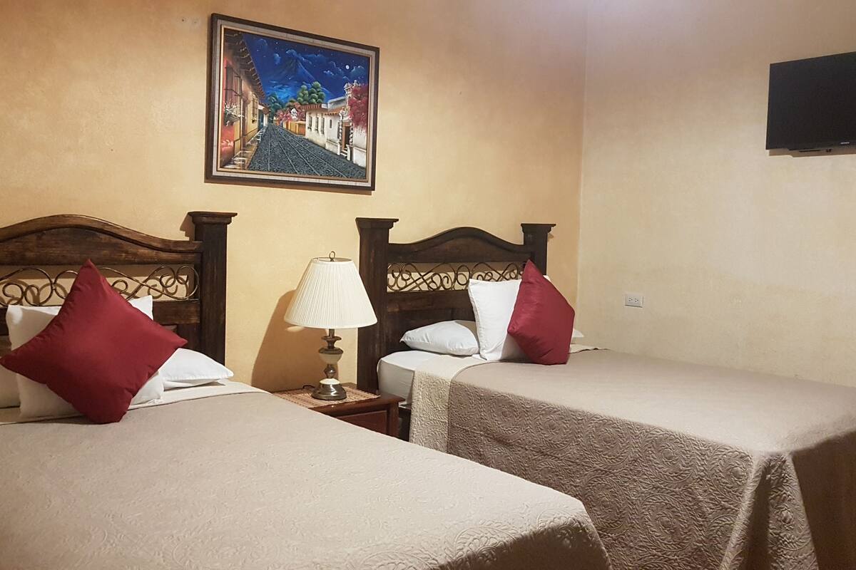 Quarto de hotel com duas camas, cada uma com cabeceira de madeira, roupa de cama branca e almofadas vermelhas. Um criado-mudo com abajur fica entre as camas. Um quadro com uma cena de rua está na parede acima das camas.