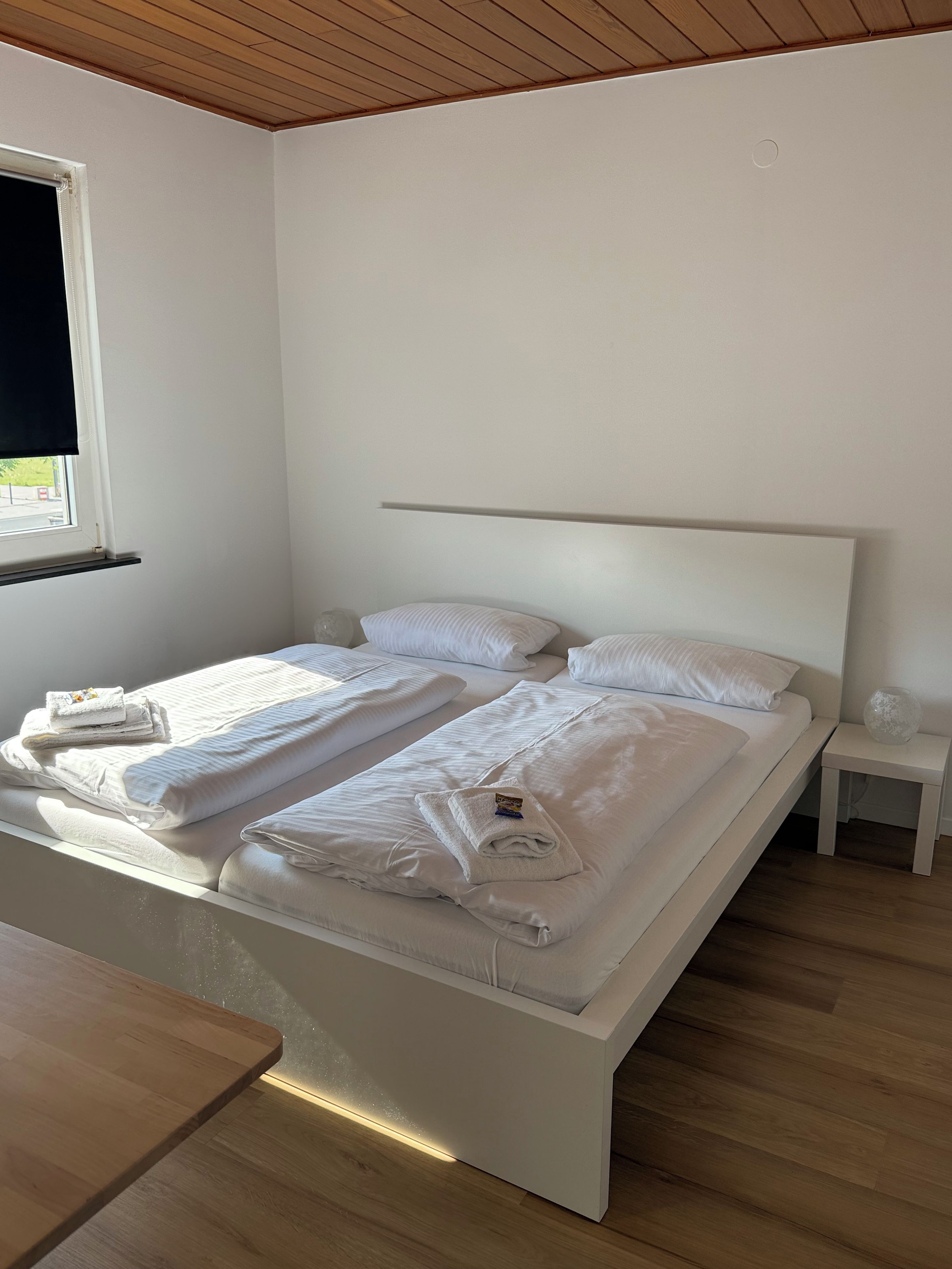 Una cama bien hecha con sábanas y toallas blancas, una mesita de noche con un florero de vidrio y una ventana con vistas al exterior.