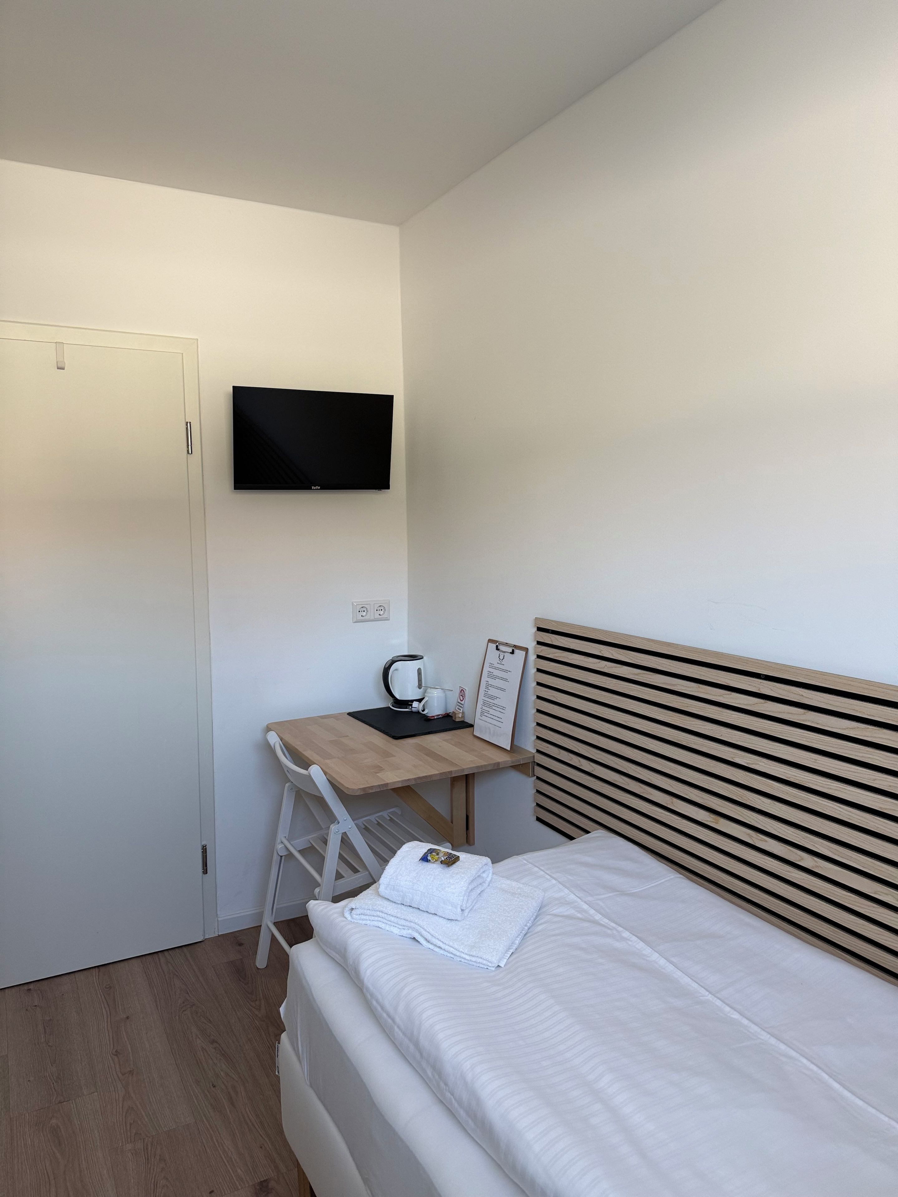 A bedroom with a bed, a desk with a kettle, a television, and a wooden headboard.