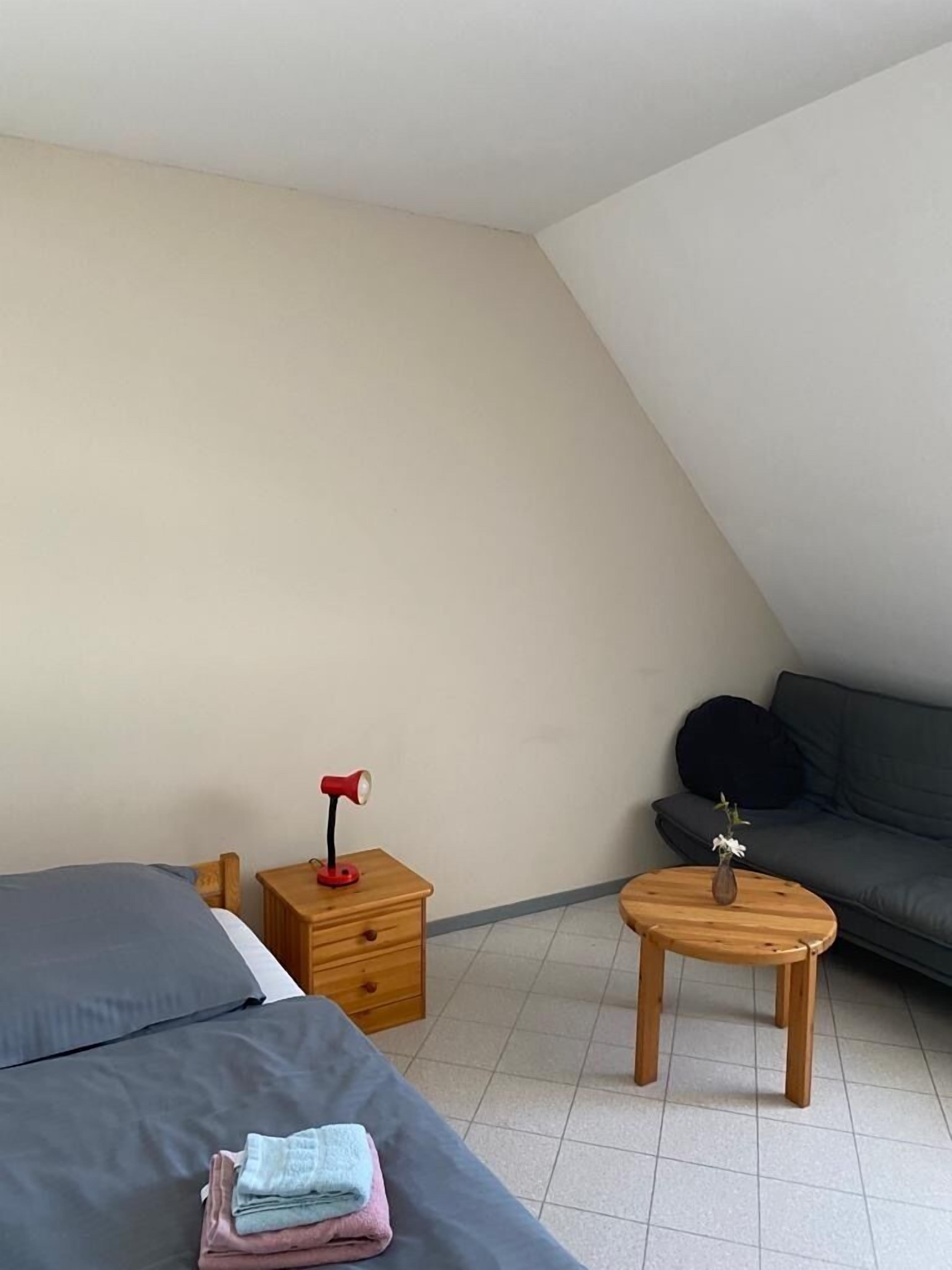 Una habitación con una cama, una mesita de noche con lámpara, una pequeña mesa con un jarrón y un sofá.