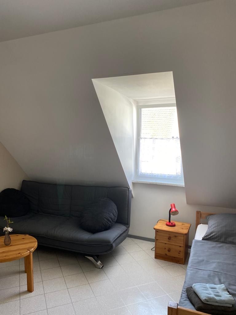 A small, well-lit room with a sloped ceiling, a sofa, a bedside table, a lamp, and a window.